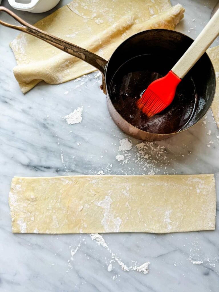 A pot of browned butter is ready to spread on a strip of puff pastry dough.