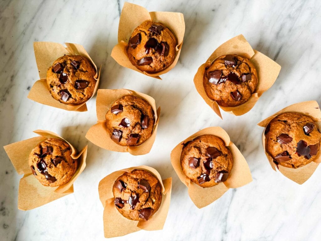 Pumpkin spice muffins with molten chocolate in the middle and dark chocolate chunks on top are in paper muffin wrappers and sitting on a kitchen counter.