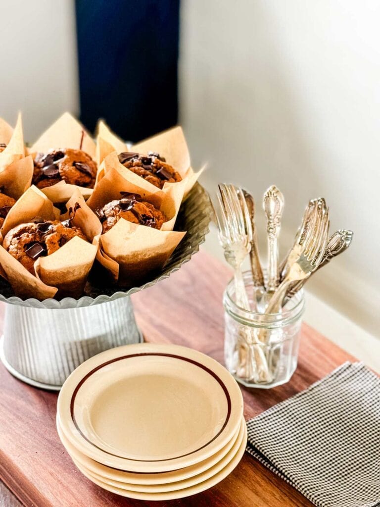 Pumpkin spice muffins with molten chocolate are on a galvanized metal cake stand next to a stack of plates and a jar with forks. Linen napkins are on the side. 