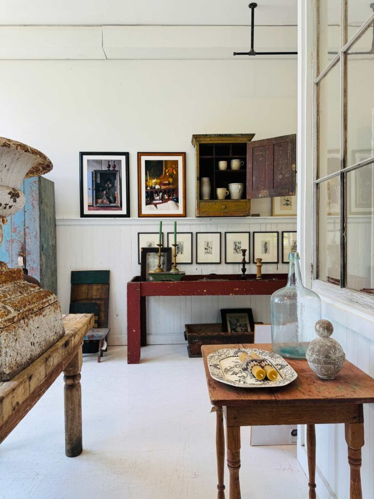 shop with art on walls, cabinet on wall, red long table with objects, antique table, platter, bottle.