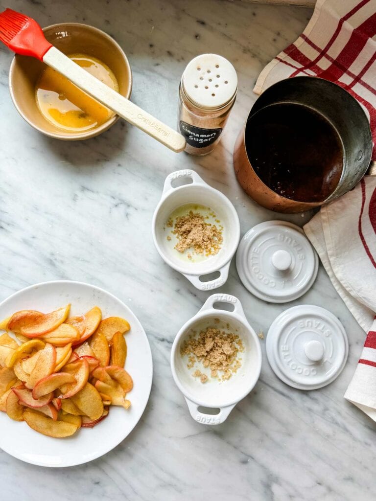 A plate of softened apples, a pot of browned butter, and mini Staub cocottes with brown sugar in the bottom are sitting on a kitchen counter.