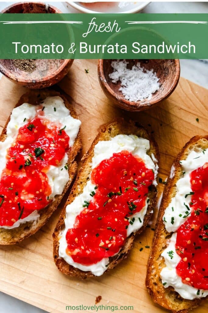 Fresh tomato and burrata sandwiches are sitting on a large wooden cutting board with containers of salt and pepper next to them.