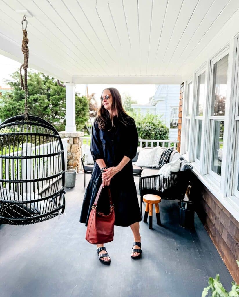 older woman wearing black dress, black sandals, brown bag on porch.