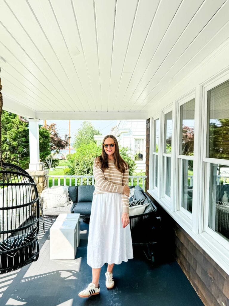 Older woman wearing a stripe cashmere sweater with a white midi skirt, sneakers on a porch.  