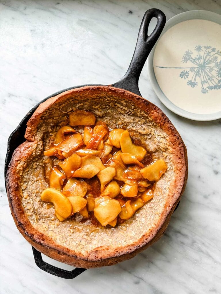 Caramelized apples have been added to the baked Dutch Baby pancake in a cast iron skillet.