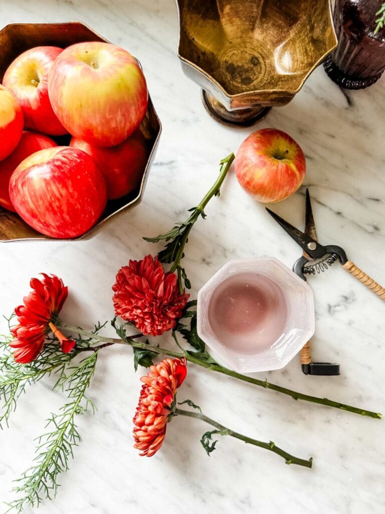 Apples in gold bowl, flowers cut on counter, pruners, pink cup/vase. 