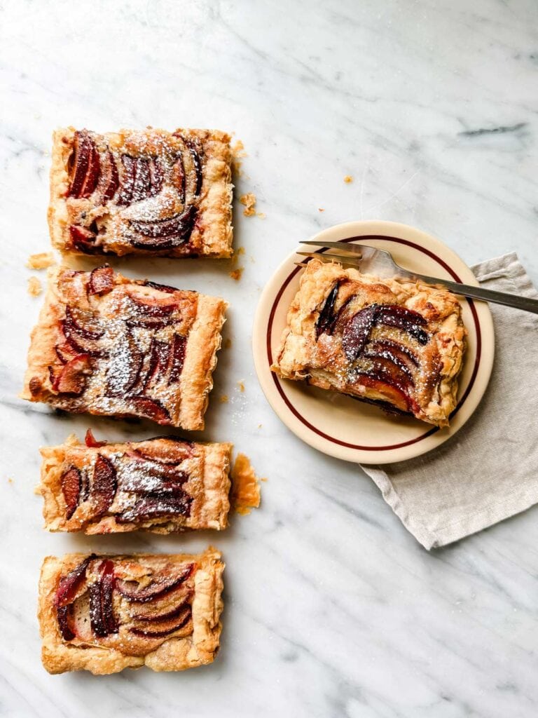 Plum and Frangipane Tart Recipe: A Seasonal Dessert. Slices of frangipane plum tarts are on the kitchen countertop and one slice is on a small brown plate ready to be served.