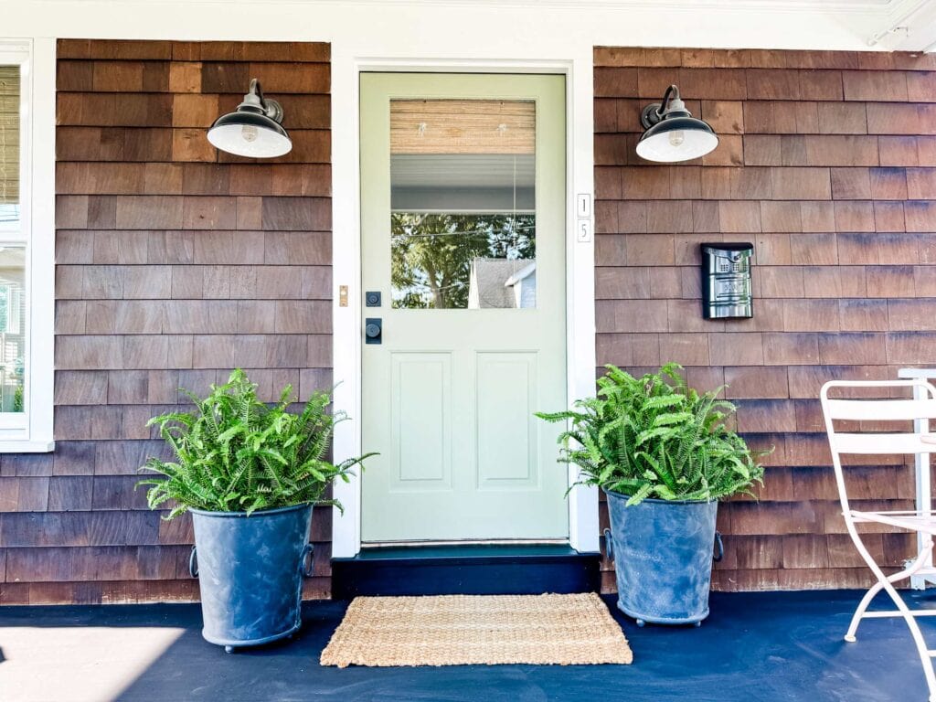 The front door of a Nantucket-style house has been painted sage green. Next to the door on each side are Ferm in metal black pots. Off to the side is a pink metal table and a pair of chairs.