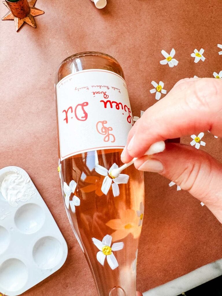 Painting daisies with yellow centers on wine bottle of rose.