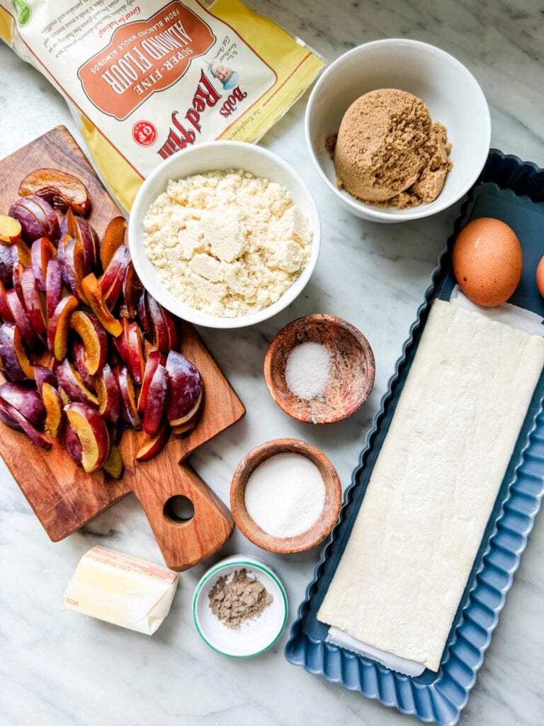 Ingredients for making frangipane plum tarts are laid out on the marbled countertop ready to be assembled and baked.