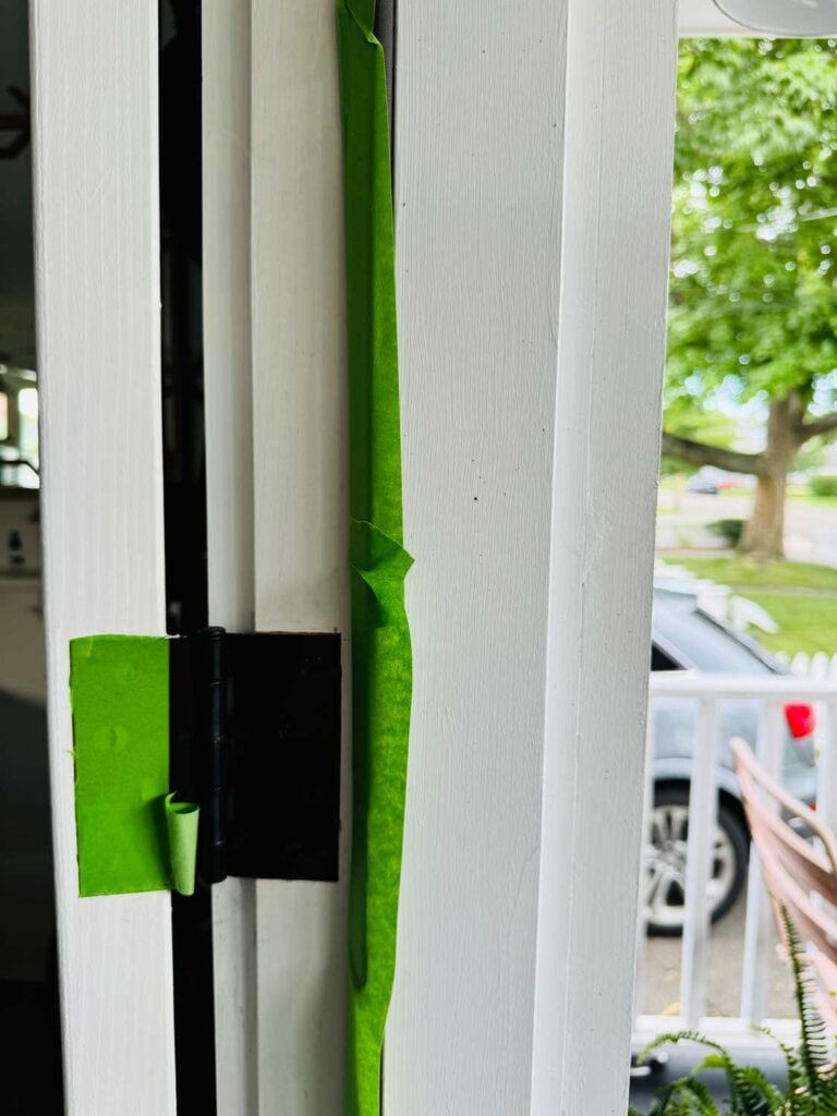 The hinges and weather stripping of a front door have been prepared with painter's tape and are ready for painting.