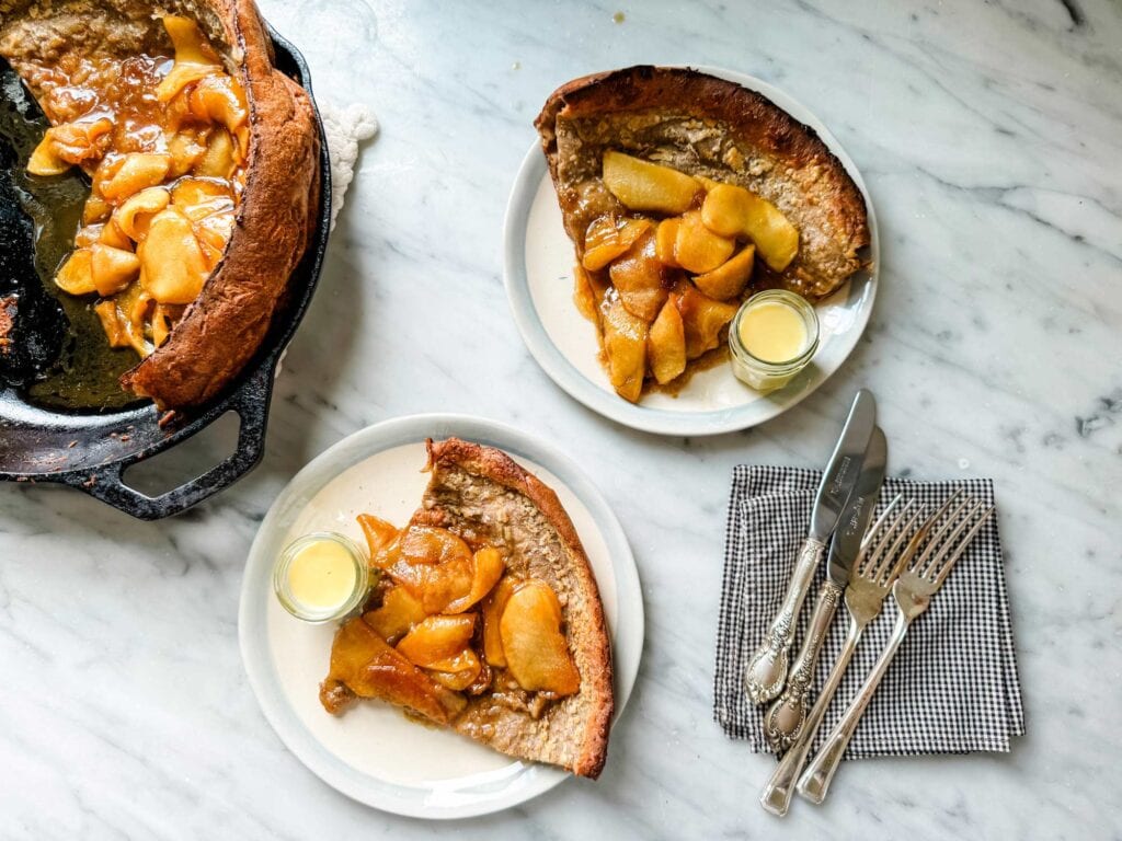 A Dutch Baby pancake is in a cast iron skillet and two pieces have been cut and placed on small plates with a jar of crème anglaise. Knives and forks are place on napkins beside the plates.