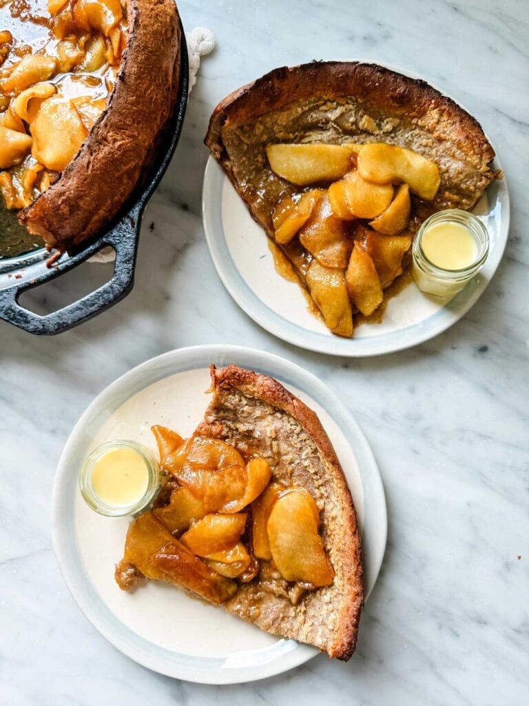 A Dutch baby pancake with caramelized apples is in a cast iron skillet. Two slices are severed on small plates with Bonne Maman jam jars filled with crème anglaise.