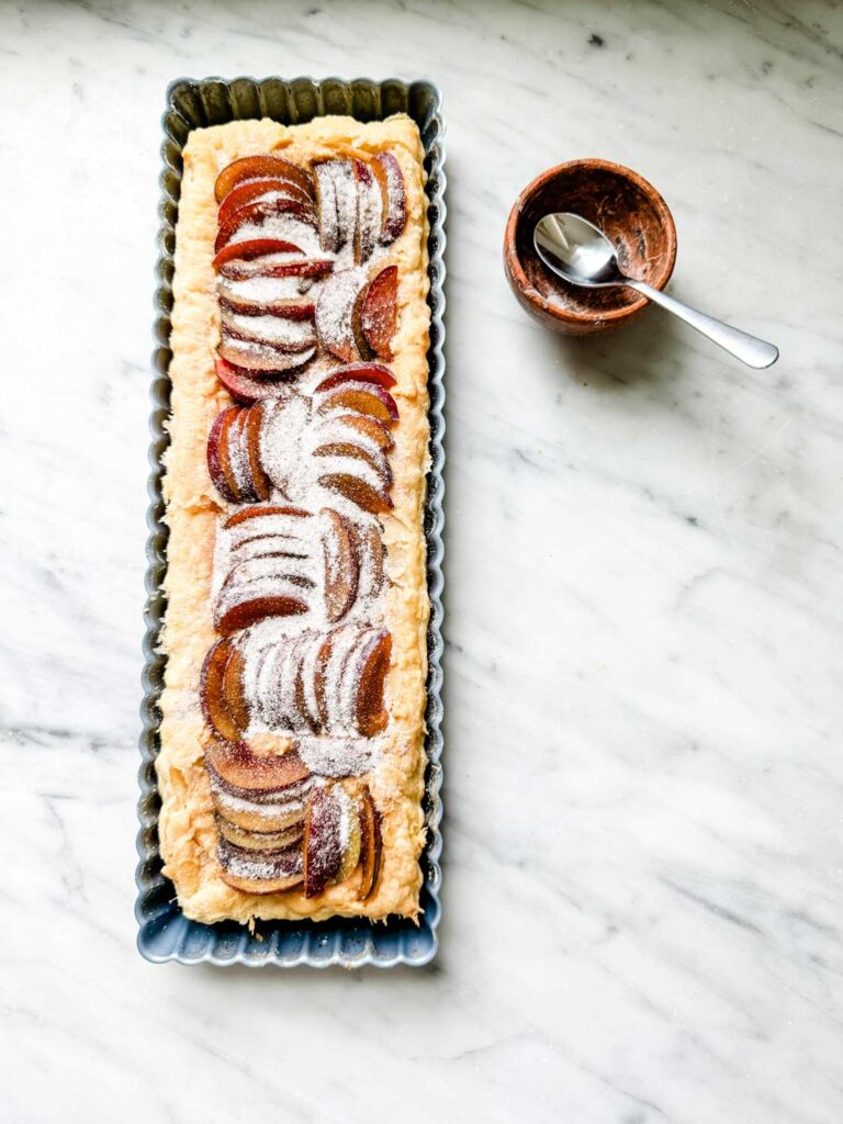 rectangular tart tin with puff pastry and uncooked plum slices arranged in rows, sprinkled with sugar. 