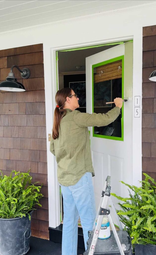 A woman is painting the front door of a Nantucket-style house. A can of paint is sitting on a step stool.