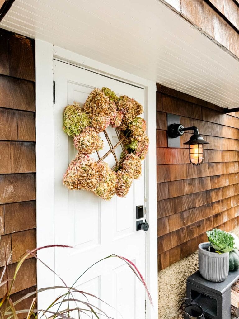 A rustic fall wreath is hanging on the side door of a house. Next to the door is a Barn Yard Electric light. A bench with pots is in the background.