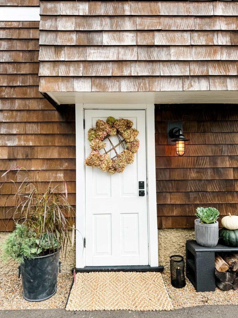 On the side door of a house is a rustic hydrangea wreath is used to decorate for fall. Next to the door are various pots filled with fall grasses and plants.