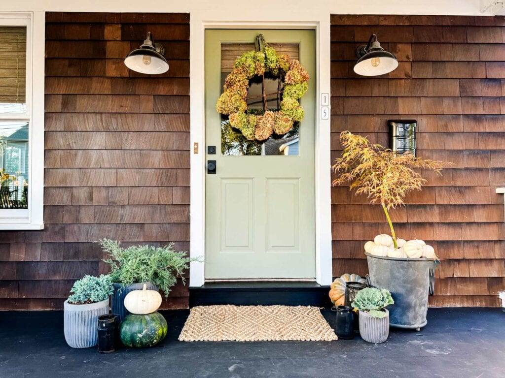 A rustic hydrangea wreath without a wreath form is hanging on the front door of the house. The front door is green, the house is brown, and the wood floor of the porch is stained black.