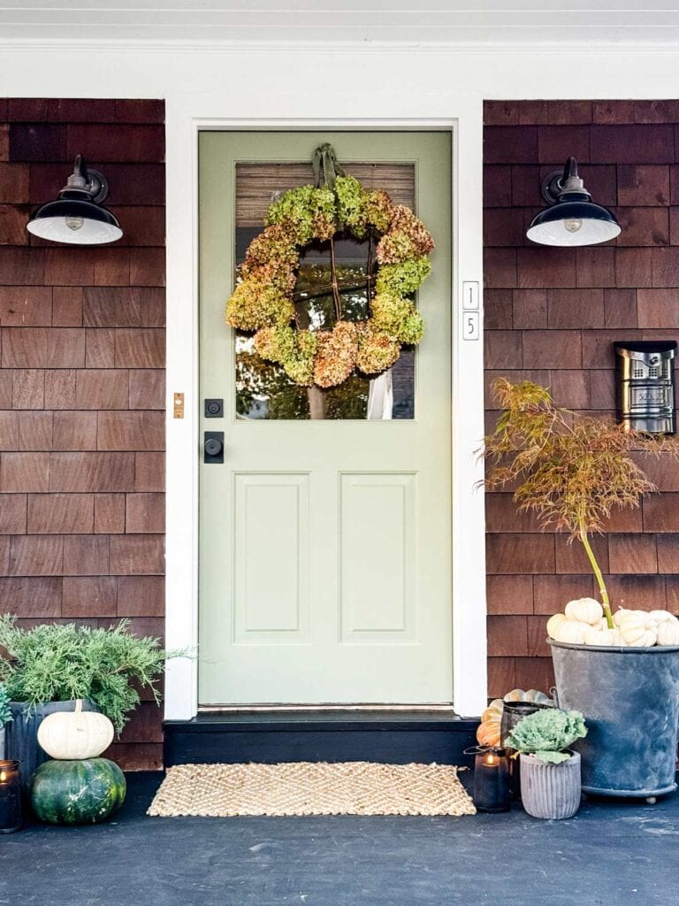 Two lights are next to the front door of a house and in the glass frame hangs a rustic hydrangea wreath.