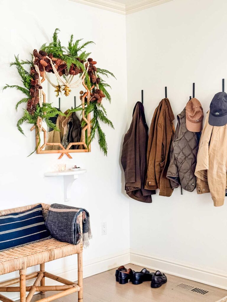 Faux garland is hanging around a rectangular mirror. Next to the mirror is a coat racking with hanging coats and hats.