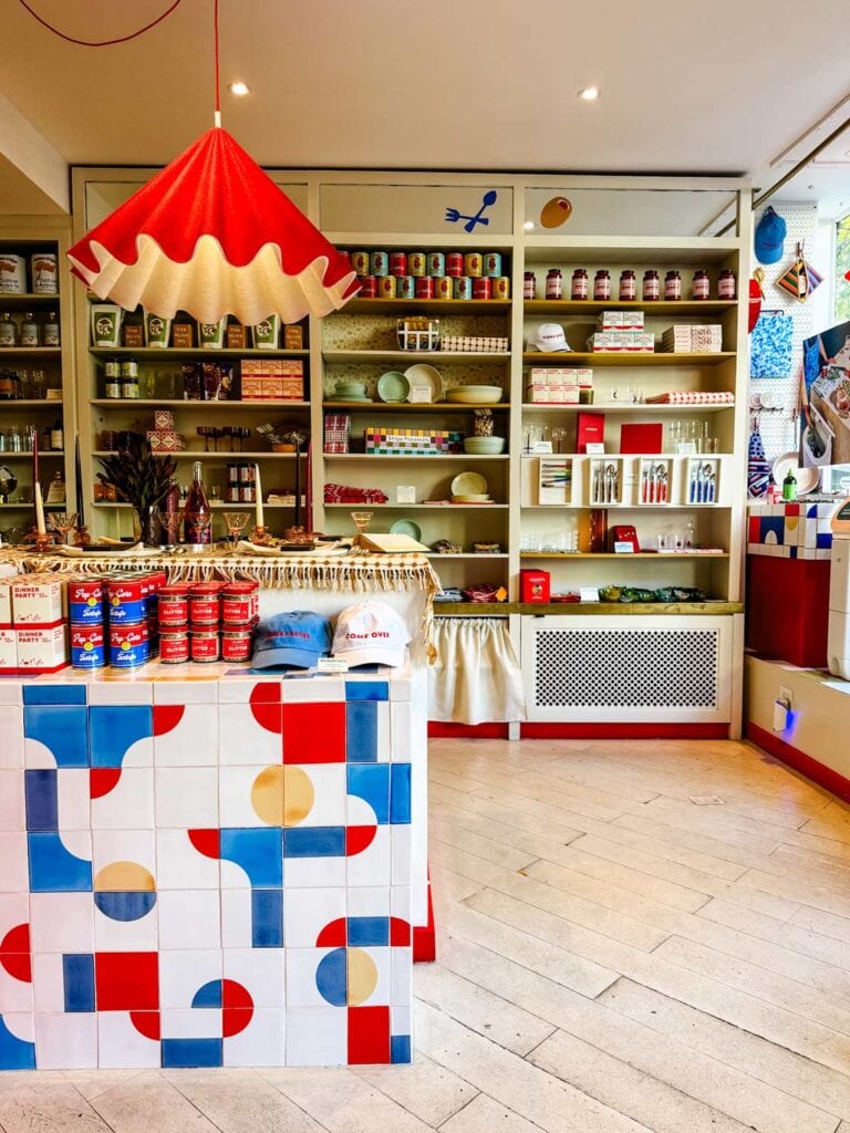 The shelves at Big Night in the Westi Village of NYC are full of all kinds of food. In the foreground is a red, white, blue and yellow counter with geometric shapes. On top of the counter are jars and cans of food along with a few hats.