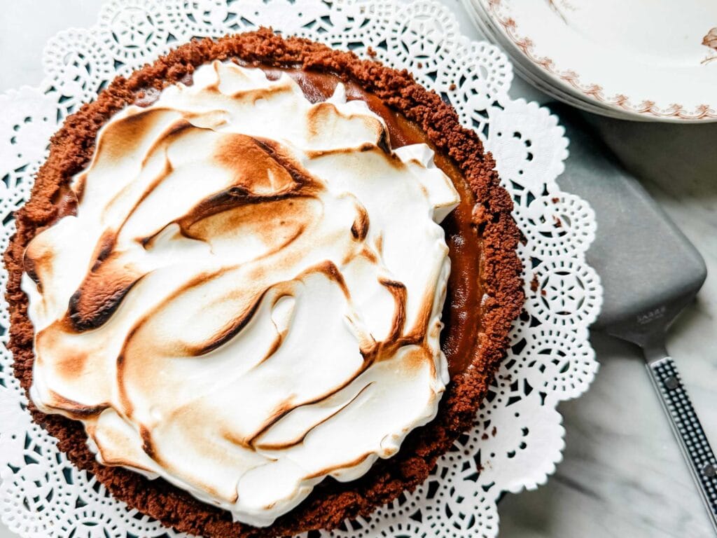 A pumpkin pie with toasted meringue is on a plate with a doily. Next to the plate is a serving spatula.