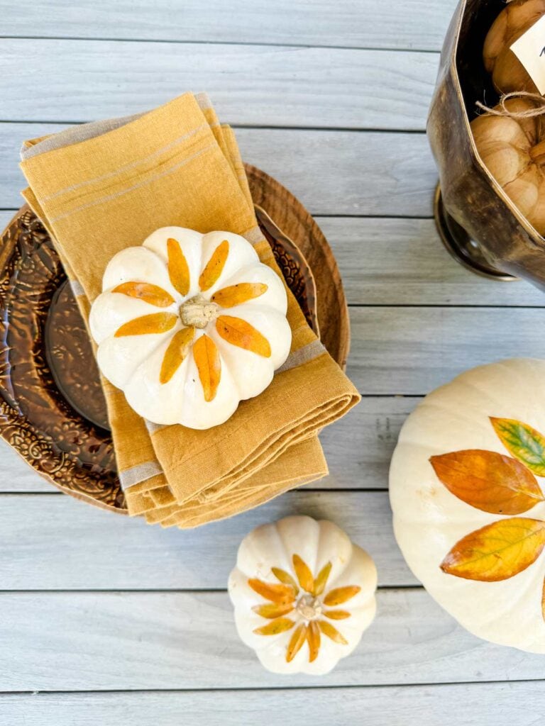 White pumpkins, fall leaves, on kraft paper bag. Brown plates with gold linen napkins. 