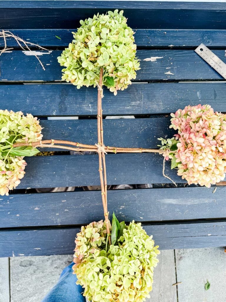 Hydrangea stems have been tied together to for a cross. They are on a black potting bench.