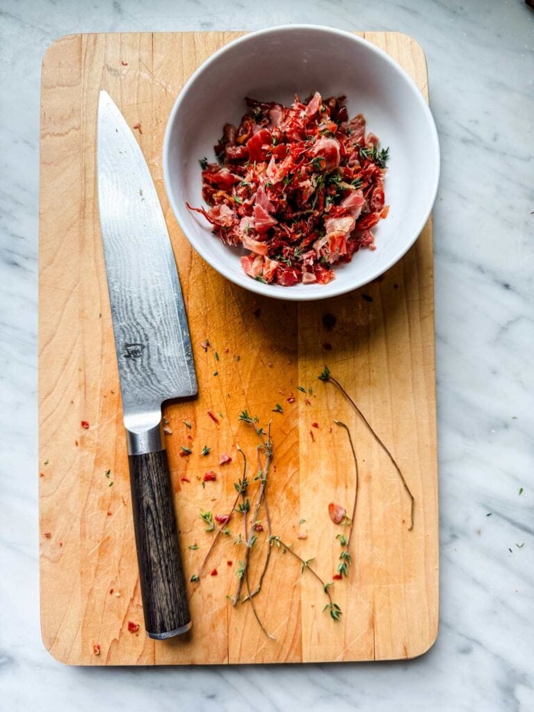 Chopped crispy prosciutto mixed with fresh thyme is in a small white bowl.