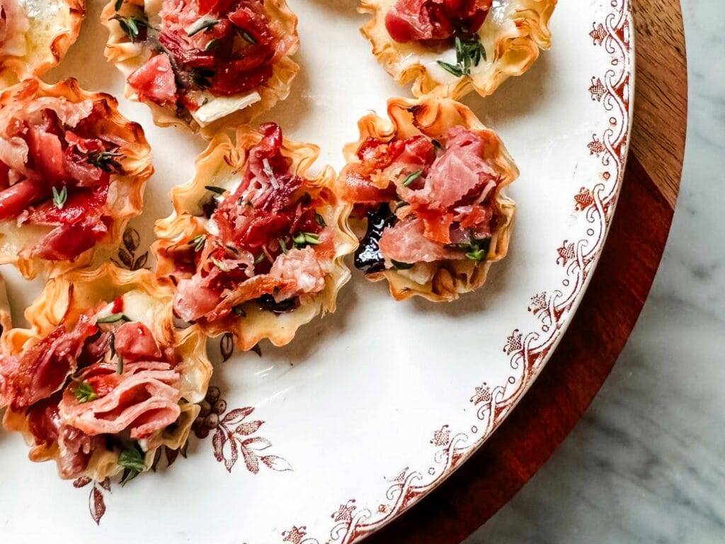 Crispy prosciutto and brie appetizers in mini phyllo cups are on a serving plate.