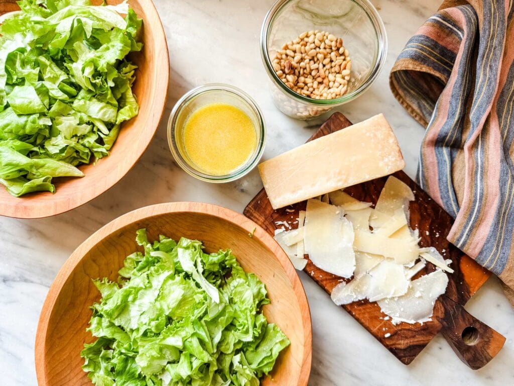 Green salads in wooden bowls next to parmesan cheese on a cutting board, a Weck jar of the vinaigrette, and a jar of toasted pine nuts.