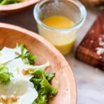 A jar of healthy and delicious Meyer lemon vinaigrette salad dressing is next to two wooden bowls filled with fresh green salad.