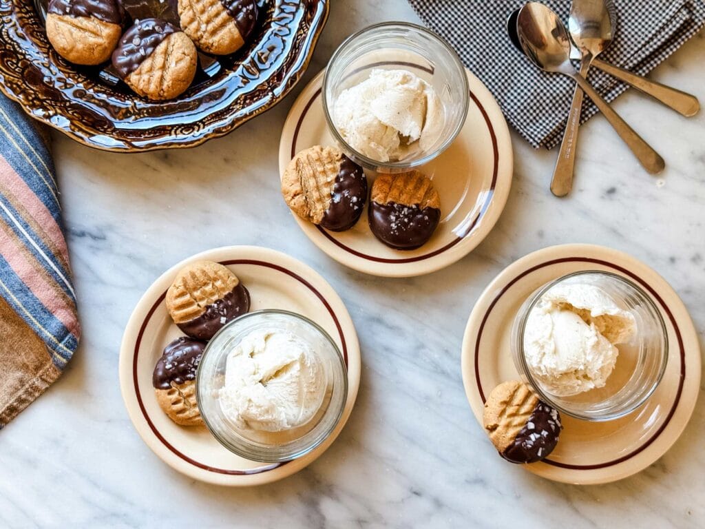 Peanut butter cookies half dipped in chocolate on small plates with cups of vanilla gelato.