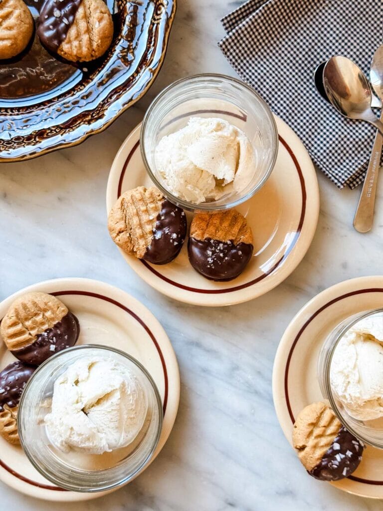 Peanut butter cookies half dipped in chocolate on small plates with cups of vanilla gelato.