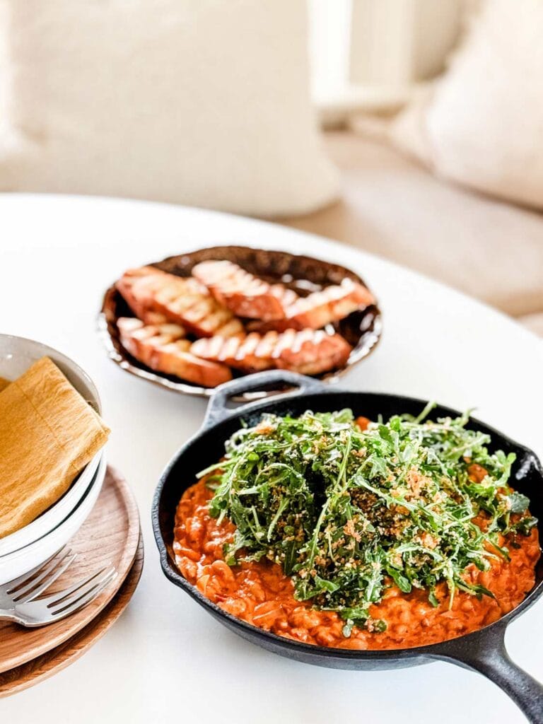 A white, round table is set for a cozy dinner for two that includes spicy tomato beans topped with arugula and served with warm toasted bread.