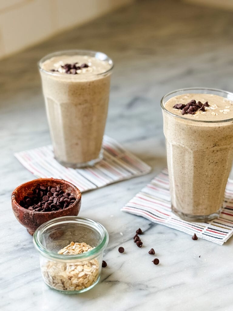 Two healthy date protein shakes are served in clear glasses and topped with oats and mini chocolate chips.