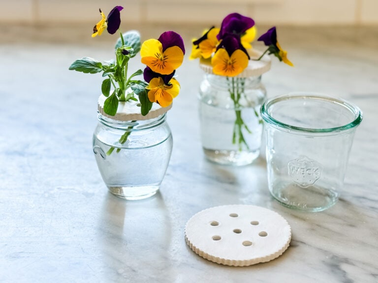 Two small glass jars have a flower frog on top, and yellow and black pansy stems are inserted into the flower frogs.