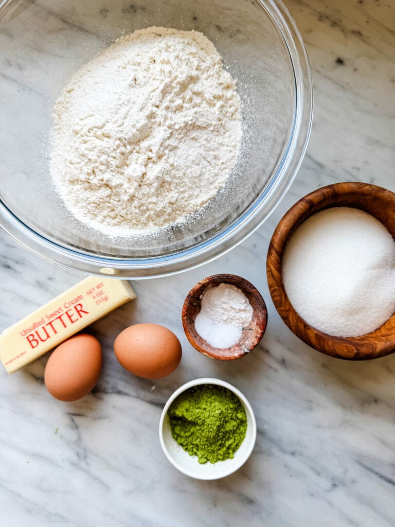 All the ingredients for making matcha crinkle cookies are measured, placed in containers, and are sitting on a kitchen counter.