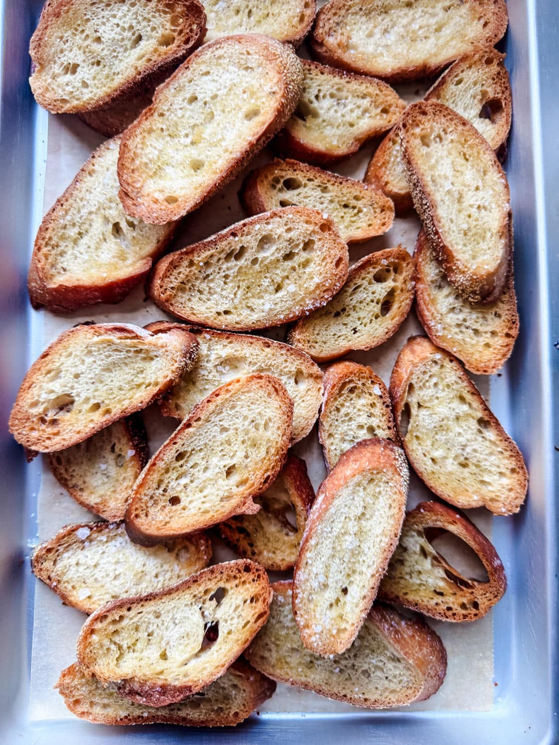 Baguette slices haven been roasted to make crostini for baked goat cheese, tomato, and olive spread.