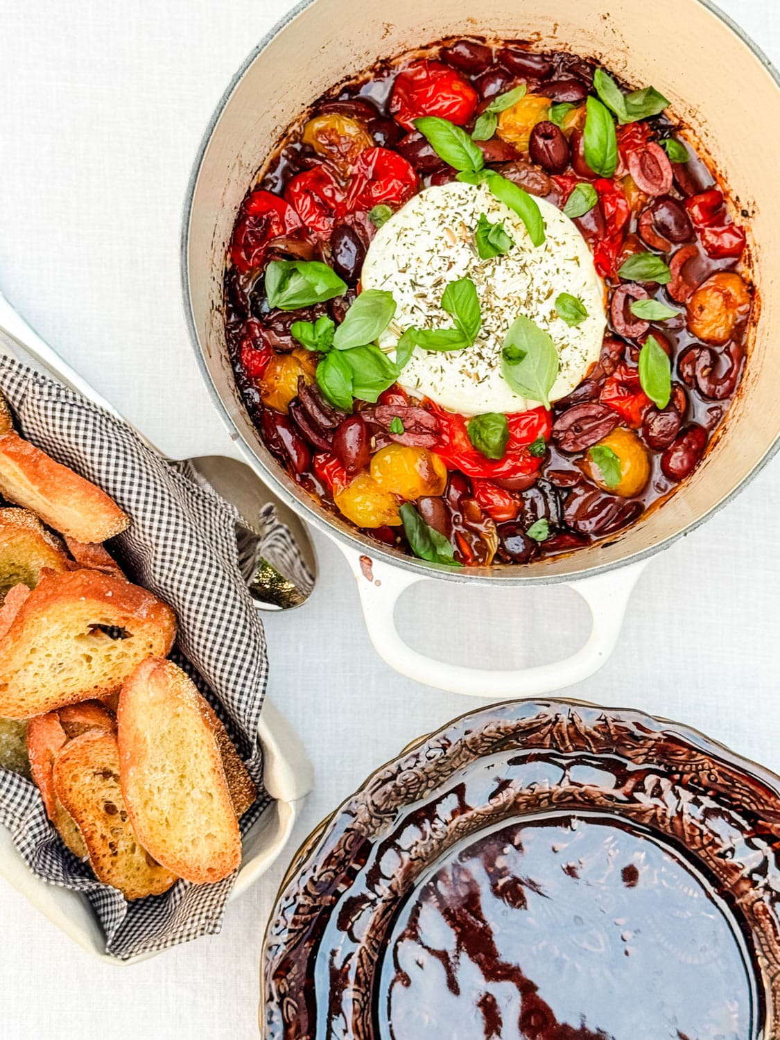 A Dutch oven with baked goat cheese spread next to crostini and a stack of appetizer plates.