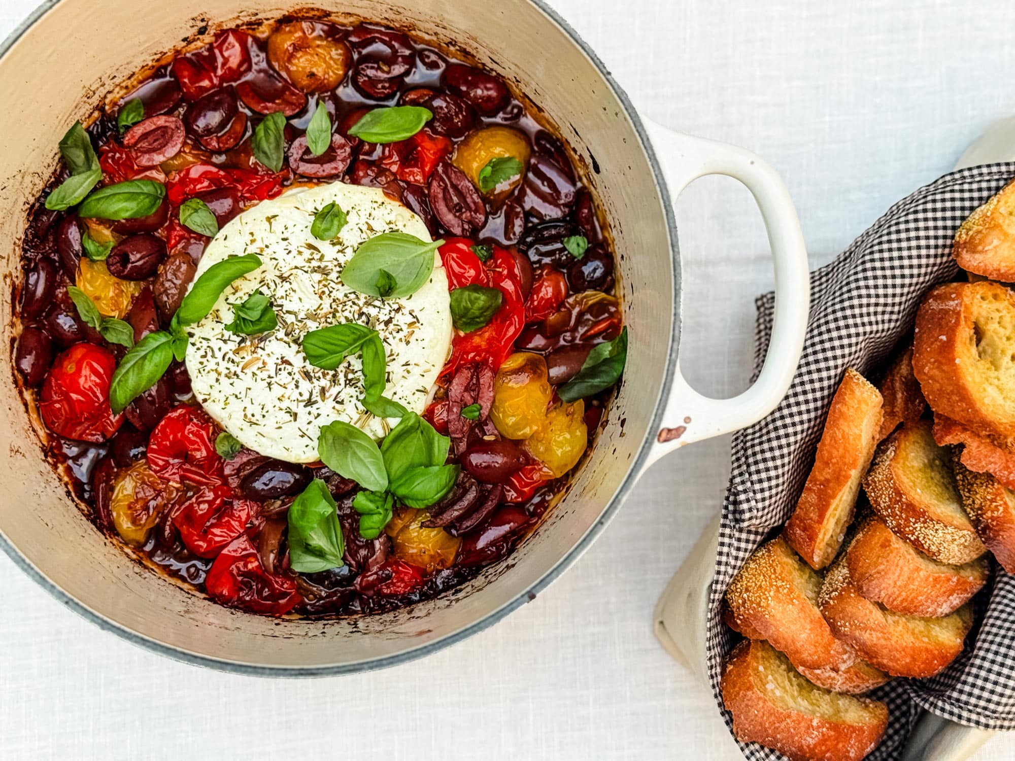 Baked goat cheese, tomato, and olive spread in a Le Cruseutte pan and served with crostini in a small, linen-lined bread basket.
