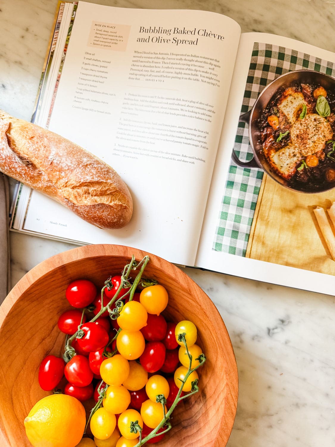 The Mostly French cookbook is open to a recipe. Next to the books is a baguette and a wooden bowl full of red and yellow tomatoes and a lemon.