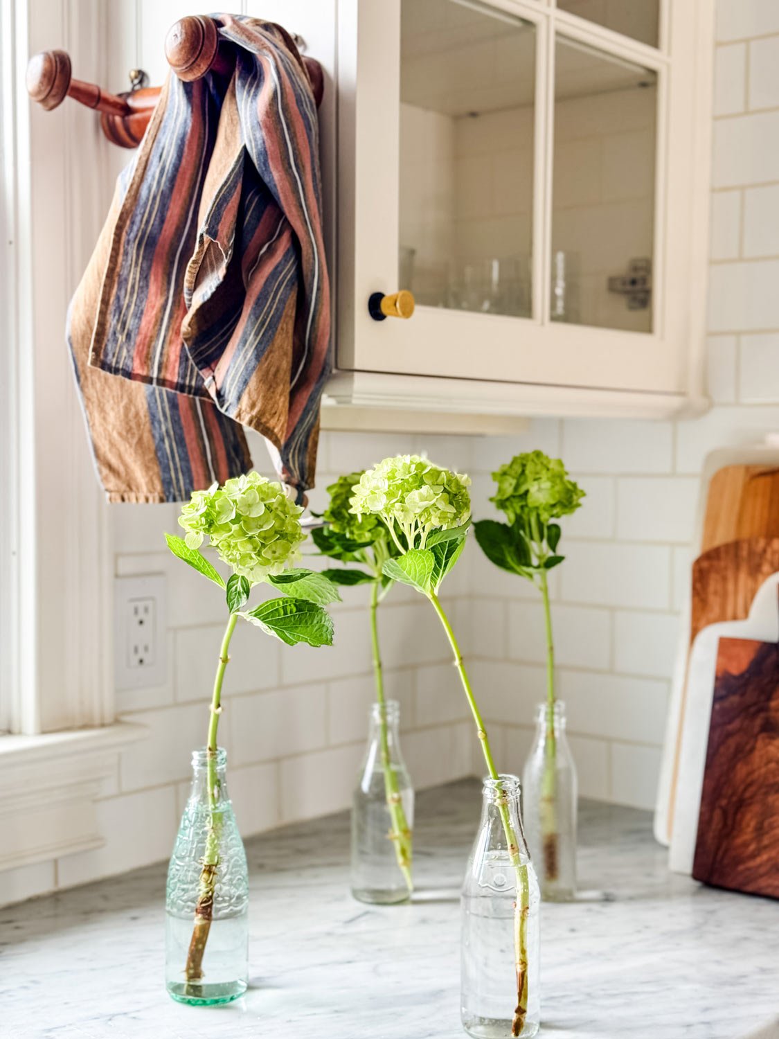 Hydrangeas in bottles on marble counter with stripe dish towel on french peg rack.