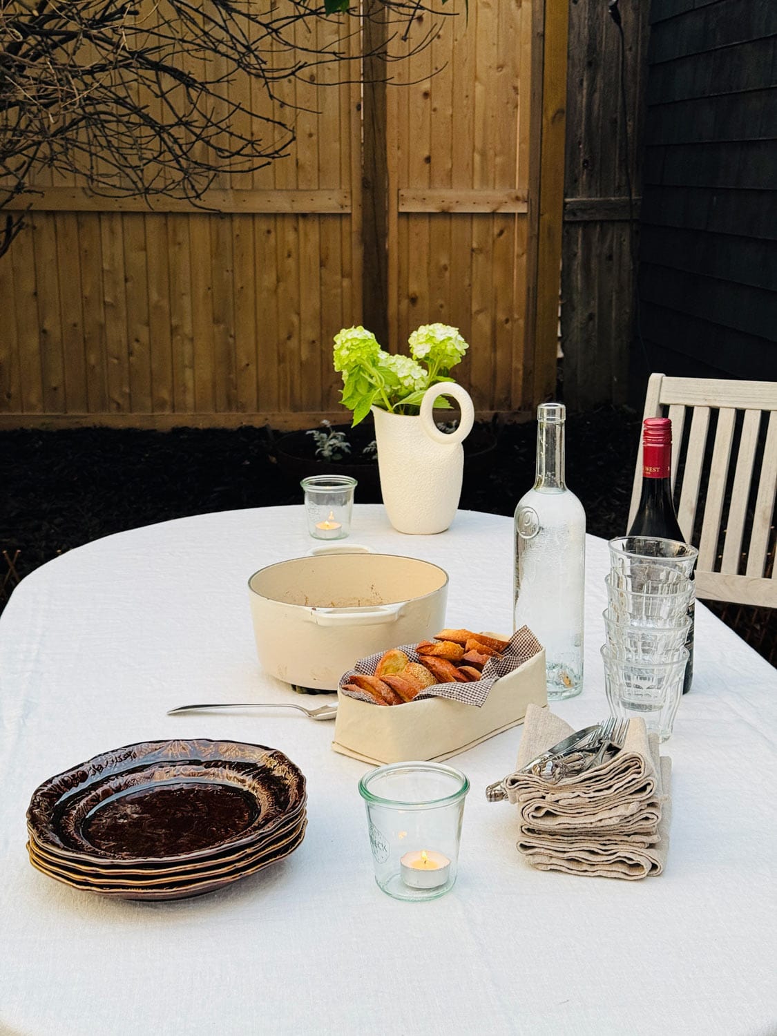 An outdoor table is set with glasses, water, wine, plates, napkins, crostini, and baked goat cheese spread.