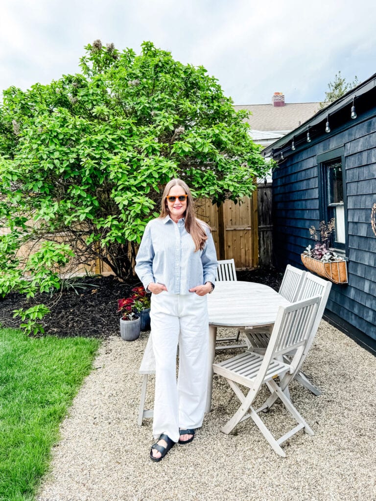 Woman wearing white jeans and striped shirt in a garden with an outdoor table and chairs.