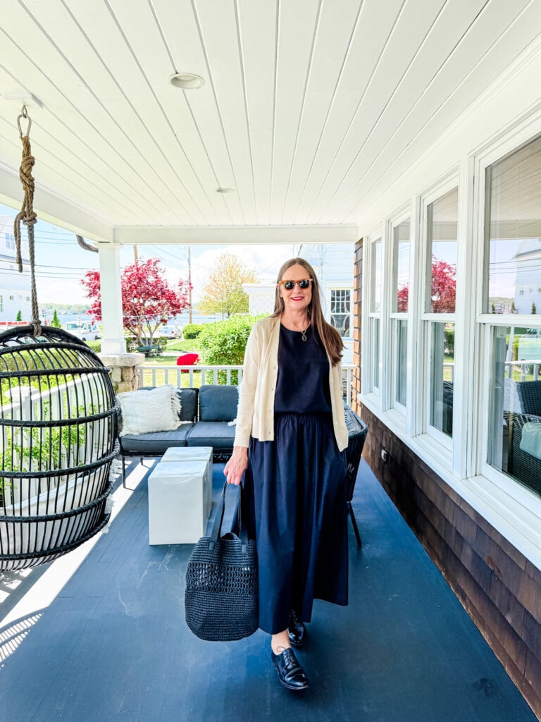 Woman wearing black skirt, tank, sweater with black bag on a porch.