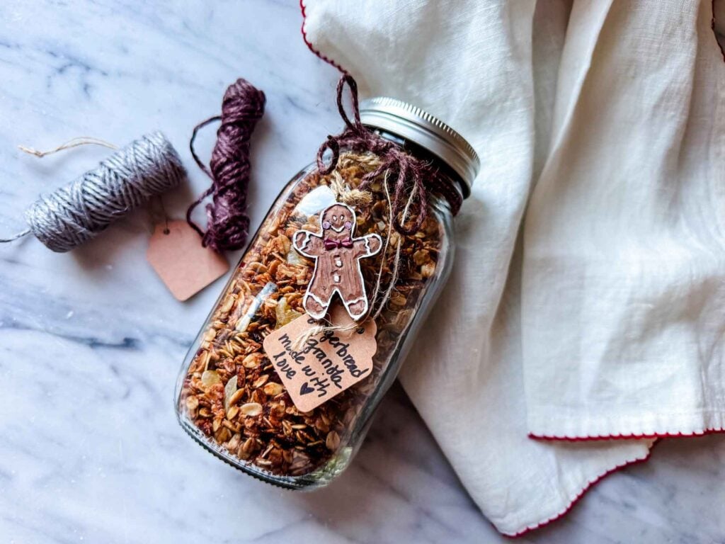 Granola in clear glass jar on marble counter with twine, dish tower with a red border.