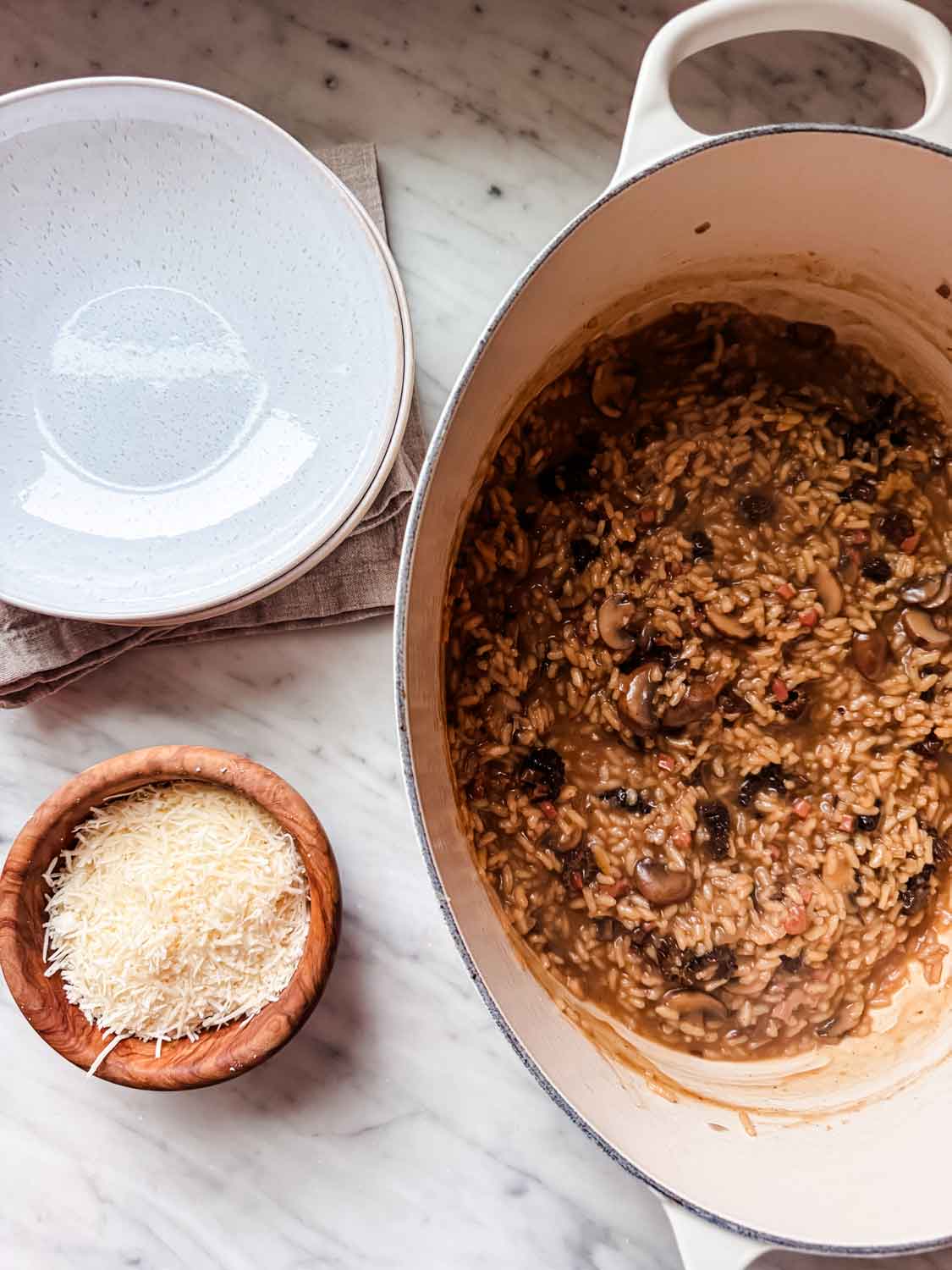 Mushroom risotto is cooked in a large Dutch oven. Bowls for serving and freshly grated Parmesan cheese are sitting next to it.