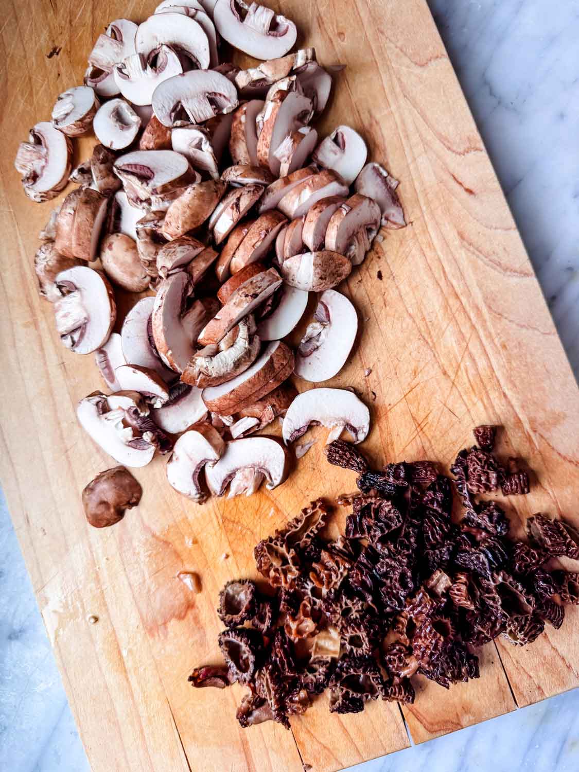 Slice baby bella mushrooms and chopped morel mushrooms on a wooden cutting board.