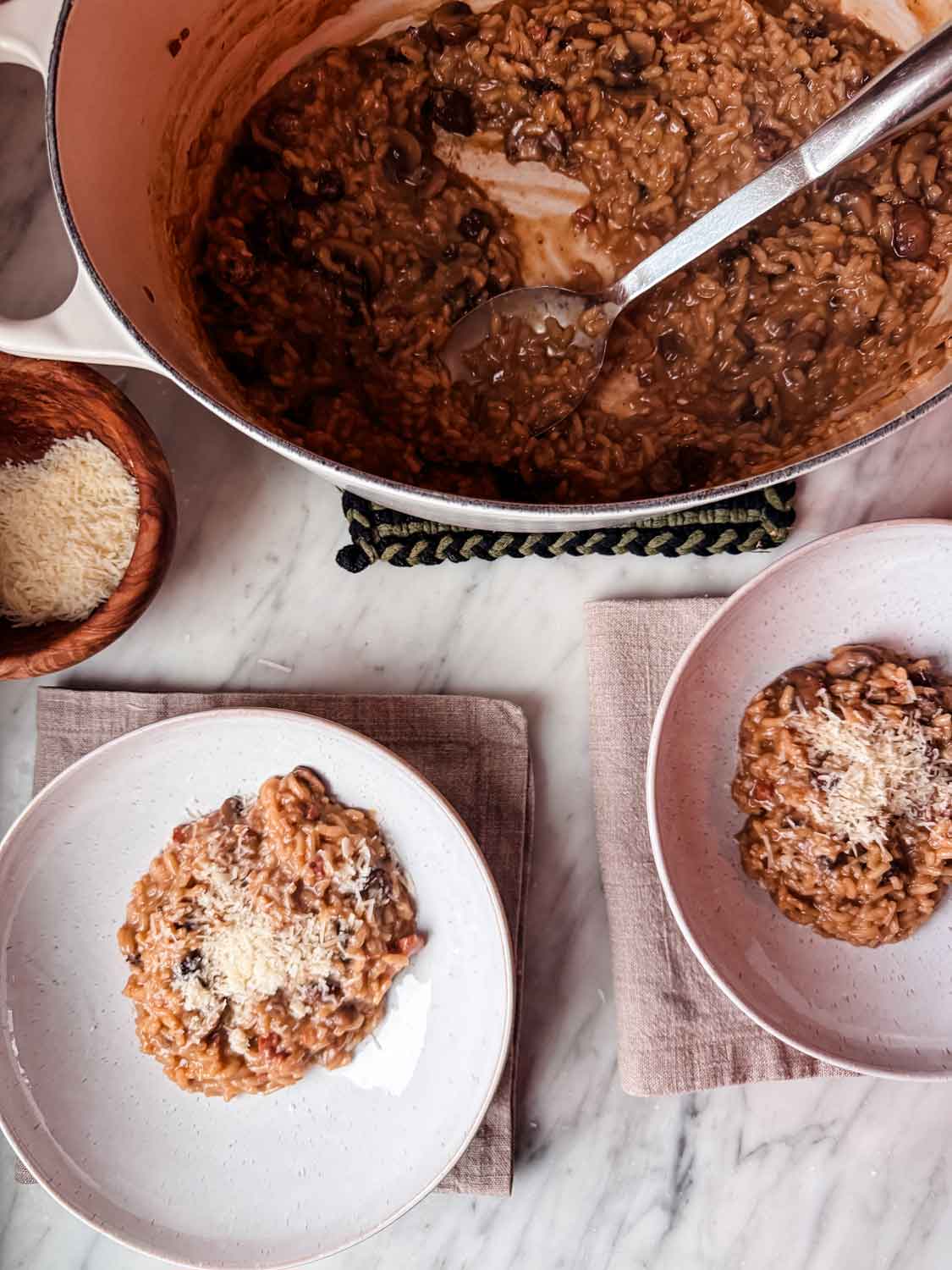 Mushroom risotto is in a large Dutch oven, cooked, next to two individual bowls with risotto and topped with parmesan cheese.